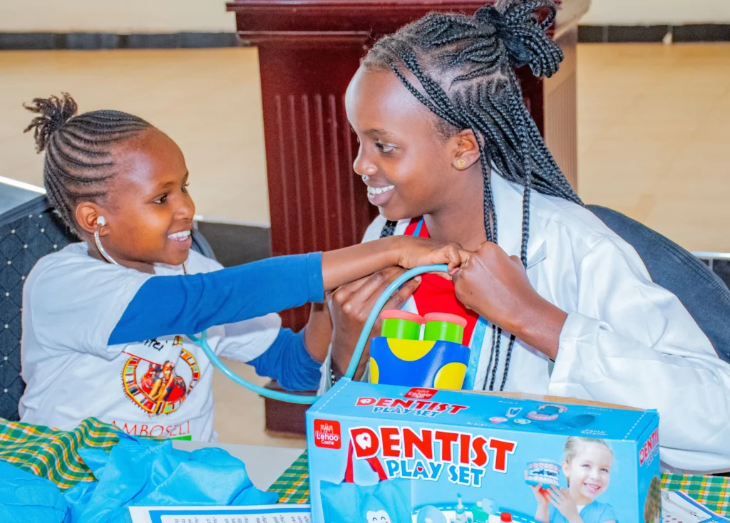Two young girls smile while playing with a Dentist Play Set, one holding a stethoscope to the other's chest during an educational activity.