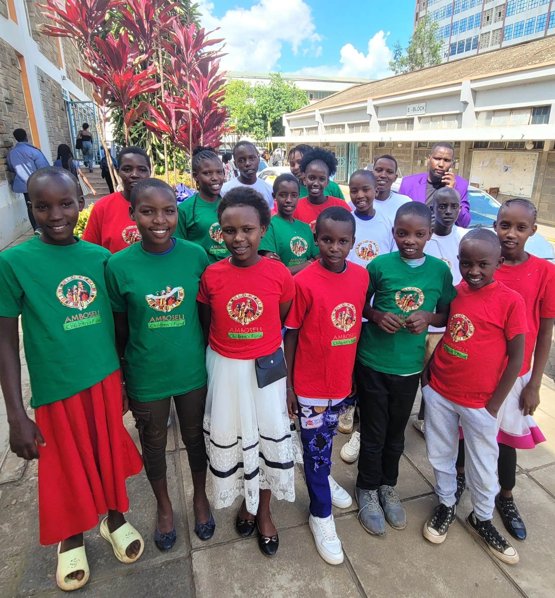 Group of children wearing red and green Amboseli Children's Fund t-shirts posing together in a courtyard.
