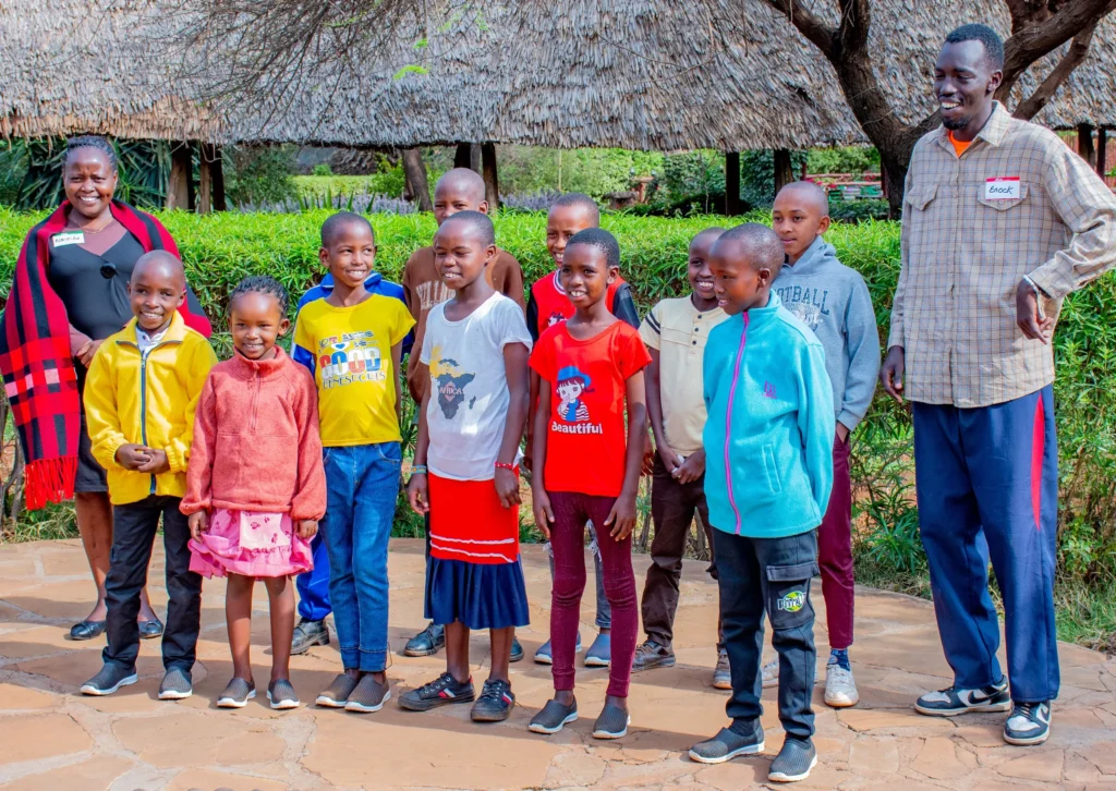 Group of smiling children and two adults standing together outside a thatched-roof building with lush green vegetation in the background.