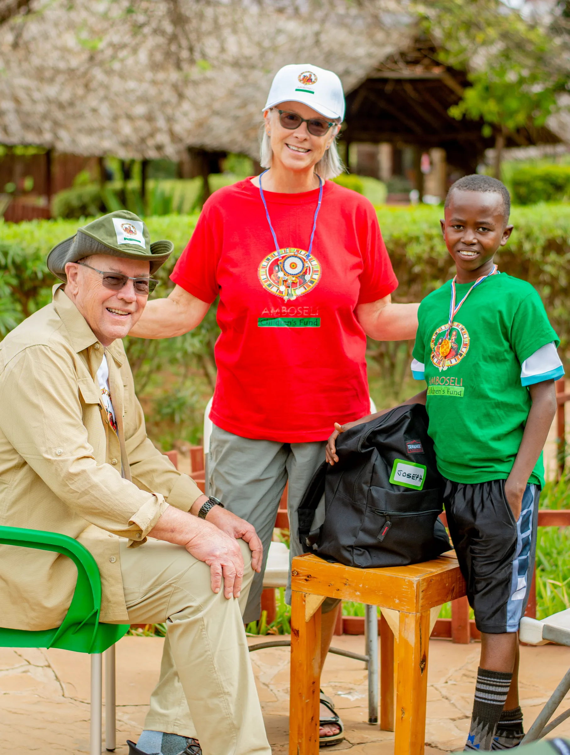 Three people outdoors at Amboseli Children's Fund: two smiling adults in logo shirts and caps